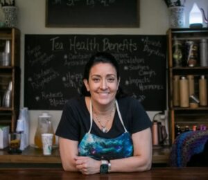 Angel Altman, nurse and owner of Tea of Life Apothecary in Chillicothe, Ohio, leaning over the apothecary counter surrounded by herbal teas and natural wellness products.